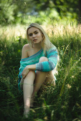 Gorgeous woman with attractive legs sitting on ground in park. Shot of girl outside in forest wearing only knitted jumper sitting in grass and looking into camera.