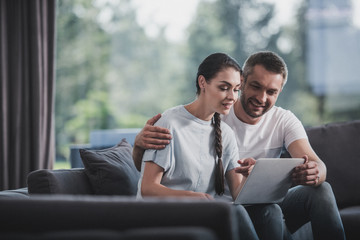 happy man embracing girlfriend and teaching her using laptop on couch at home