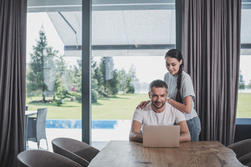 smiling young woman embracing boyfriend while he using laptop at home