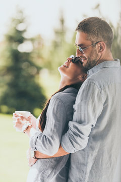 side view of happy man in sunglasses embracing girlfriend with coffee cup outdoors