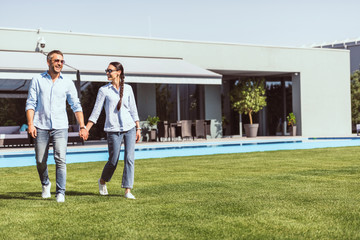 smiling couple holding hands and walking on green lawn near swimming pool at country house