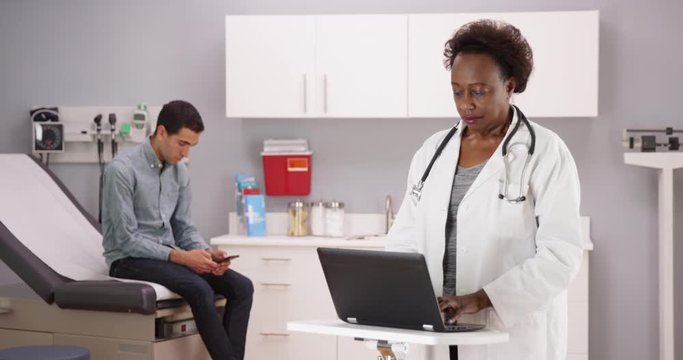 Wide shot of medical clinic with mid aged African doctor typing on laptop while young male patient uses smartphone. Senior black doctor taking notes of single latino patient on portable computer