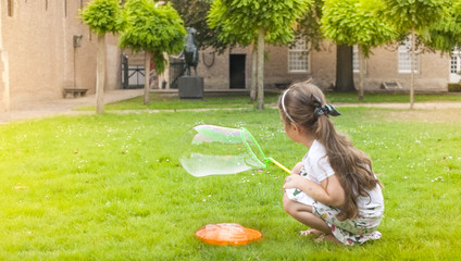 Fototapeta na wymiar Little girl is playing with soap bubbles in a park on green grass. Happy childhood and outdoor games. Free space for text. Copy space.