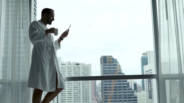 Happy man in bathrobe using tablet computer and drinking tea by window at home
