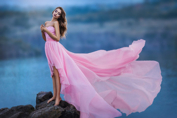 Gorgeous woman stands on the rock in pink tender dress. Flying cloth like wings