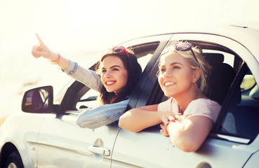 summer vacation, holidays, travel, road trip and people concept - happy teenage girls or young women sitting in car at seaside and pointing finger to something