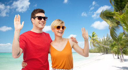 travel, tourism and summer holidays concept - smiling couple in sunglasses hugging and waving hands over exotic tropical beach with palm trees background