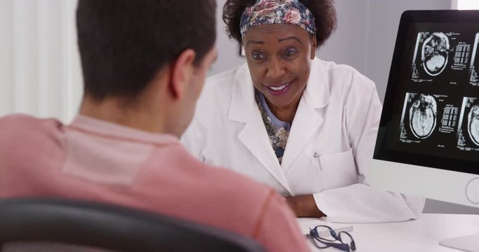 Middle aged black doctor consulting with young male patient about ct-scans of his cranium. Young male patient looking at scans of his brain on computer at doctors office