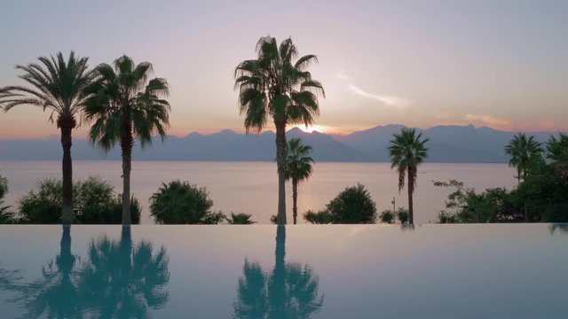 Evening scene with outdoor swimming pool with palms overlooking the sea and sun going down behind the mountains, Turkey