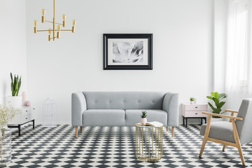 Grey couch and armchair on checkered floor in white flat interior with gold lamp and poster. Real...