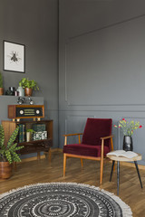 Real photo of a red armchair standing between a stool with flowers and shelf with books, radio and plants in retro living room interior