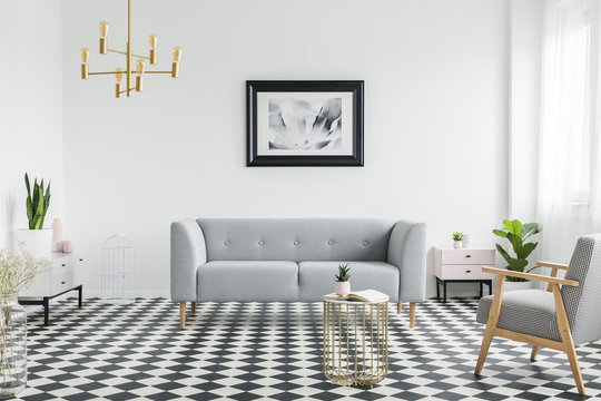 Grey couch and armchair on checkered floor in white flat interior with gold lamp and poster. Real photo