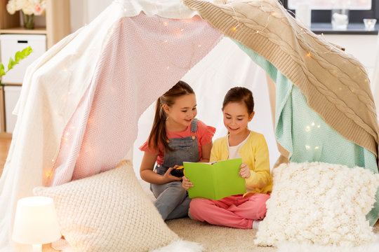childhood, friendship and hygge concept - happy little girls reading book with torch light in kids tent or teepee at home