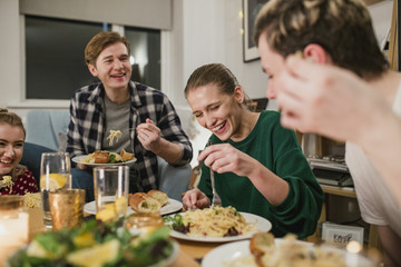 Friends are having a dinner party at home. They are laughing while eating spaghetti carbonara.