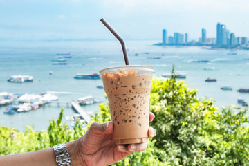 Hand holding iced coffee and sea with many boats at Pattaya in Thailand.