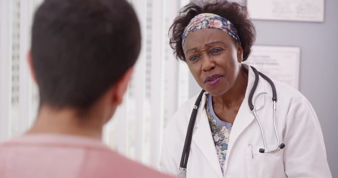 Positive female African doctor talking to male patient inside medical office. Close up of senior black MD smiling with young adult male patient and talking about health concerns