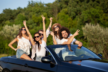 Glad stylish young dark-haired girls and guys in sunglasses smile in a black cabriolet on the road...