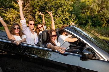 Group of young girls and guys are holding their hands up and making selfie in a black cabriolet on...