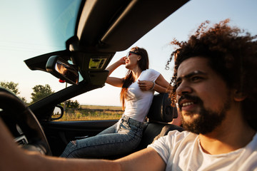 Stylish company of young girls and guys are sitting and smiling in a black cabriolet on the country road on a sunny day.