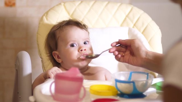 Small baby eats porridge from spoon sitting on chair in kitchen.