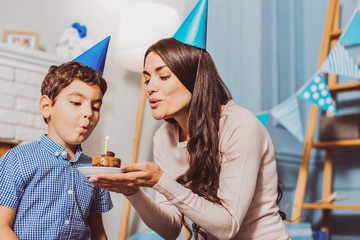 Happy birthday dear. Low angle of jovial positive woman carrying plate with birthday cake while boy extinguishing candle