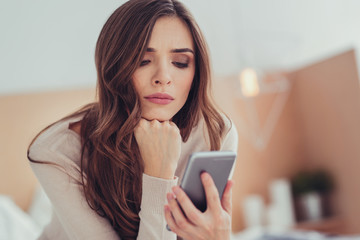 Online. Portrait of thoughtful young girl holding mobile phone and propping her face with a fist while feeling worried and waiting for messages from her boyfriend - obrazy, fototapety, plakaty