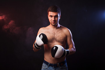 Studio portrait of a muscular boxer in professional gloves of European appearance with light bristles and hair on his chest. Smoke in the background is illuminated in blue and red