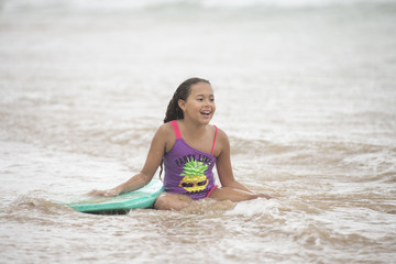 Cute young mixed race child sitting on the waters edge laughing holding a boogie board and being splashed by the waves