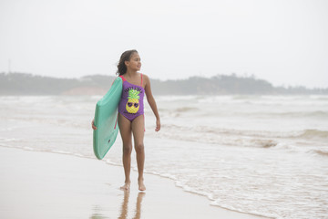 Mixed-race female child walking towards the camera on the beach and waves on a bright tropical summer holiday with boogie board under arm