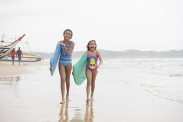 Two beautiful young mixed race sister girls walking on the beach wearing swim wear laughing with boogie boards