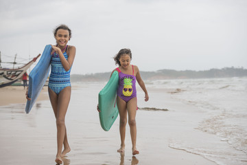 Two beautiful young mixed race sister girls walking on the beach wearing swim wear laughing with boogie boards