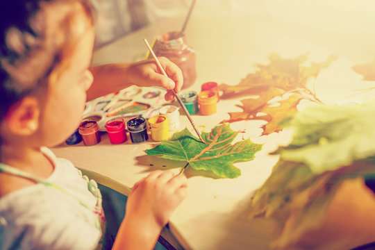 Cute little girl drawing with paint autumn leaves at home