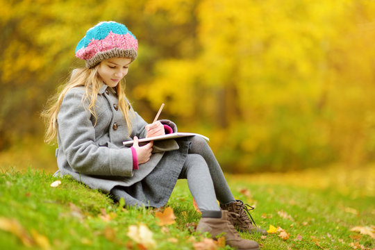 Cute little girl sketching outside on beautiful autumn day. Happy child playing in autumn park. Kid drawing with colourful pencils.