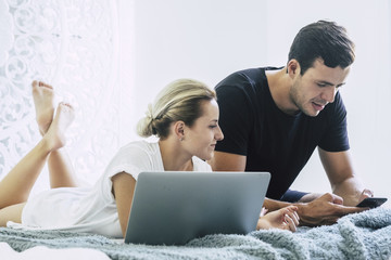 beautiful caucasian couple laying on the bed at home in the bedroom working and useing technology notebook laptop and mobile phone. enjoying  the modern activity together working on devices