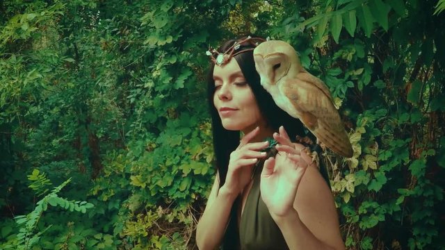 A forest nymph with long dark hair walks with a white owl. A girl in an unusual green dress with a deep neckline and bare legs. Background wild nature