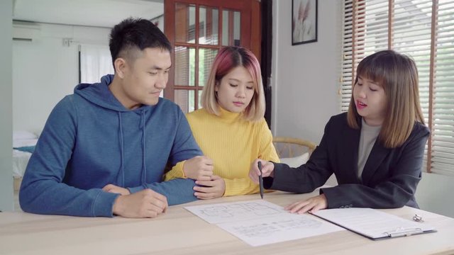 Happy young Asian couple and realtor agent. Cheerful young man signing some documents while sitting at desk together with his wife. Buying new house real estate. Signing good condition contract.