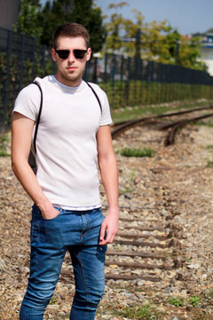Handsome young guy with sunglasses posing and enjoying a nice sunny day. Urban city environment. Old railway in background. Male model photo shoot, urban fashion, lifestyle. White T-shirt and jeans.