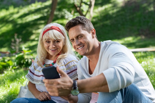 Stylish adulthood. Joyful senior woman sitting near her partner while staring at modern gadget
