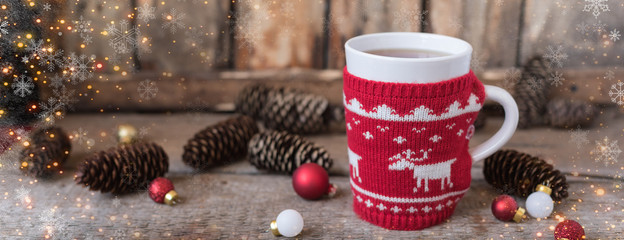 Knitted red cup with white reindeer, christmas lights with cones and toys at rustic background,...