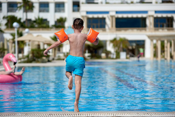 European boy in floating sleeves running to jump into swimming pool at resort.