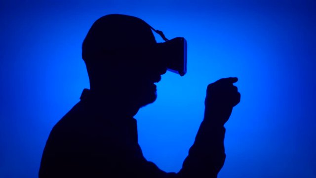 Black contour silhouette of senior man using VR 360 glasses make gestures with hands. Male's face in profile on blue background. Seeing something exciting, learning to use contemporary technologies
