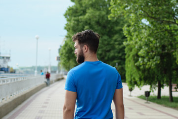 a handsome young man with a beard stands with his back to us and looks into the distance