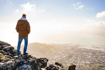 Deurstickers Tafelberg Young man is sitting on rock at Table Mountain and looking at Cape Town, South Africa