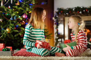 Happy little sisters wearing Christmas pajamas playing by a fireplace in a cozy dark living room on Christmas eve.