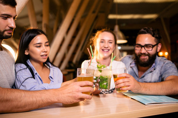 Four cheerful friends with alcoholic drinks clink glasses sitting at a table in the bar