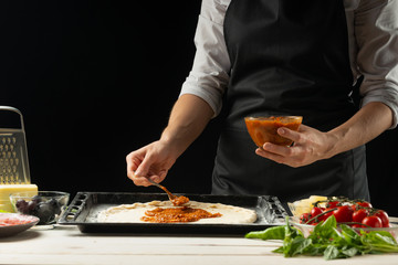 Raw pizza ingredients on a wooden table, the chef prepares pizza on a dark background