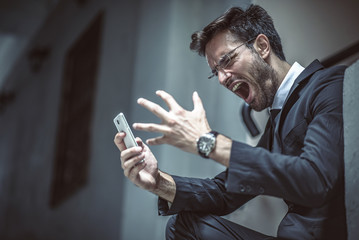 Angry, furious business man shouting at his cell phone, sitting outside a building