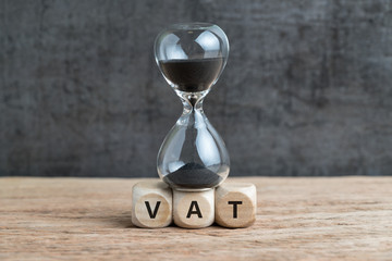 Time countdown for VAT increase deadline concept, hourglass or sandglass on cube wooden block with alphabets VAT, Value Added Tax on wood table with dark black loft cement wall
