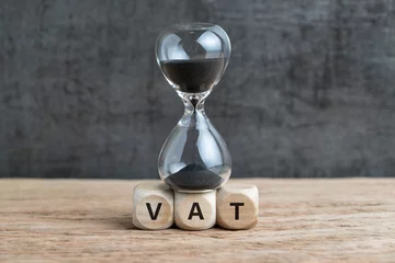 Fotobehang Time countdown for VAT increase deadline concept, hourglass or sandglass on cube wooden block with alphabets VAT, Value Added Tax on wood table with dark black loft cement wall © Nuthawut