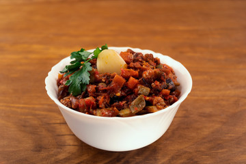 Vegetable stew with a slice of garlic and a sprig of parsley in a deep white bowl on a wooden background.